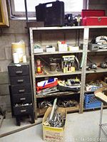 Wide shot of metal shelving unit containing multiple boxes and items including vintage blow torch, paint supplies, and tools. Black multi-drawer cabinet nearby stores small parts.