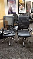Photo of two black Rolland office chairs with mesh backs and padded seats. One chair has a Logitech keyboard and mouse placed on the seat.