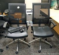 Two Rolland Whisper office chairs with mesh backs and black cushioned seats, metal bases with caster wheels, placed side by side on carpeted floor.