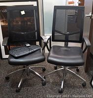Two black Rolland Whisper office chairs with mesh backs positioned side by side, one chair holds a keyboard and mouse on its seat.