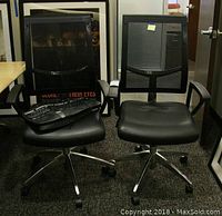 Two Rolland Whisper office chairs side-by-side. Black mesh backs, black cushioned seats, metal bases with casters. One chair has a Microsoft keyboard and microphone placed on its seat.