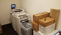 Full view of Xerox WorkCentre 7775 with attached paper trays and stacked toner boxes on top and nearby