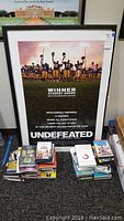Photo showing framed "Undefeated" movie poster with visible frame and glass wear, surrounded by stacks of DVDs and books on carpet floor.