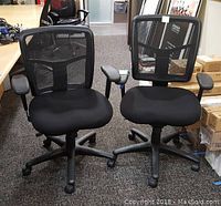Pair of black office chairs with mesh backs and fabric seats shown side by side on carpeted floor