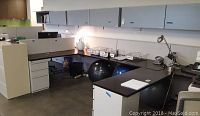 Three connected office cubicles with dark wood tops, white lower cabinets and drawers, and white overhead storage cabinets. Various office accessories and objects are on the desk surfaces.