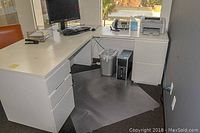 View of the L shaped white office desk setup in an office corner, featuring multiple drawers and office equipment on top.