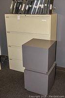 Front view of beige 4-drawer filing cabinet with multiple binders on top and two dark grey storage cubes stacked in front.