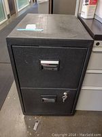 Closed front view of black metal file cabinet showing two drawers, lower drawer with lock and included key on floor.
