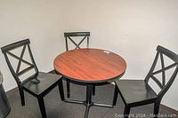 Photo showing full round dining table with wood finish top surrounded by three black wooden X-back chairs.