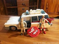 Image shows white 1980's Ghostbusters Ecto-1 replica toy car, two beige uniformed Ghostbuster figurines, and large red and white Ghostbusters logo sign on wooden surface.