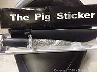 Knife in original box labeled 'The Pig Sticker'. The blade is partially wrapped in plastic and the handle is black with textured grip.