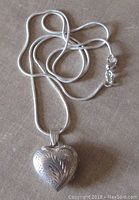 Heart shaped sterling silver engraved locket with attached chain, shown closed on a flat surface.