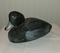 Side profile of a black wooden duck decoy with glass eyes and gray carved beak on a neutral table surface