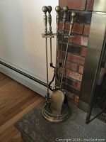 Full view of brass metal fireplace tool set with stand showing five tools arranged in circular stand on hearth in front of brick wall.
