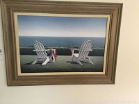 Framed oceanfront scene print by Daniel Pollera showing two Adirondack chairs with one chair draped in an American flag, framed in beige wood frame.