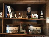 Wooden shelf with lighthouse-themed decor including ceramic miniatures, books, candle, and storage boxes