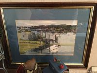 Framed seaside watercolor painting depicting boats docked by pier with bridge and buildings in background, gold frame and blue mat