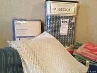 Three fabric tablecloths including one blue and white checkered cotton cloth, one blue plaid cloth, and one red cloth, along with a clear packaged vinyl tablecloth and assorted napkins.