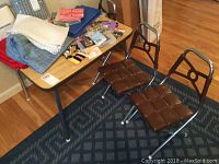 Child sized rectangular folding table with wood finish top, metal legs, three brown padded folding chairs around it. Miscellaneous items on table not included.