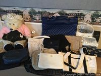 Photo showing fifteen assorted bags including Build-A-Bear Workshop plush bear wearing a princess outfit with pink sweater, tiara, and skirt; bags include quilted navy, white, cream with black trim tote, black, tan woven, silver metallic, and navy blue styles.