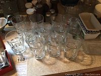 Ten clear thick-bottom juice glasses arranged on a counter among other kitchen items