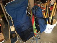 Two blue foldable camp chairs, one chair has a cup holder in the armrest, placed side by side with umbrella and bucket with baseball bats nearby.