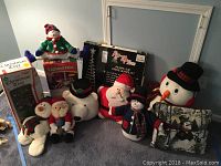 Photo showing assortment of plush Christmas Santa and snowman dolls, lighted doe decoration, boxed Christmas tree, and Christmas village pieces.
