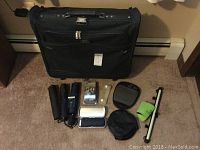 Photo showing black Olympia two-suiter rolling suitcase with 4 travel umbrellas, small mirror, green insulated sleeve, luggage straps, and other travel accessories spread out in front of the suitcase.
