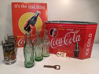 Front view of all Coca-Cola collectibles including bottles, glasses, ice chest, opener, and wall sign