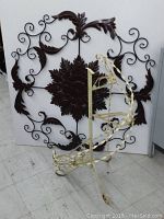 Two metal garden accent pieces against a white wall: large round dark brown leaf and scroll metal wall decoration and cream-colored wrought iron plant stand.