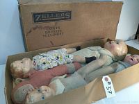 Five vintage dolls laid inside a vintage cardboard box labeled Zeller's Coats Dresses Millinery, showing the variety of the set including the largest 22 inch doll and four smaller ones.