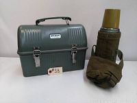 Closed vintage Stanley steel lunchbox with green color, metal clasps, and Stanley logo, alongside Stanley Aladdin thermos with original cover and attached strap.