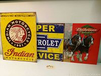 Three advertising tin signs side by side featuring Indian Motorcycle, Chevrolet, and Budweiser Clydesdales