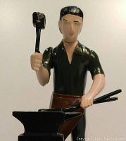 Close-up of the blacksmith figure holding hammer and tongs, painted in black, green, and brown, part of the mechanical metal bank.