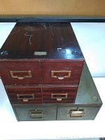 Photo showing two file drawer units stacked, one with a dark reddish finish and four brass handle drawers, and another smaller green steel two-drawer unit beneath.