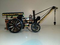 Side view of the black 1/50 scale steam toy model showing detailed wheels, crane arm with hanging weight, and yellow/red accents with company branding.