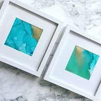 Two white‐framed abstract alcohol ink artworks displayed on marble surface