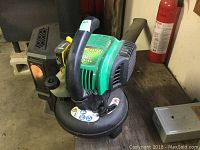 Green and black Weedeater 25cc gas blower sitting on a wooden surface in a garage or workshop environment.