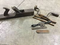 Photo showing antique wooden hand planer, hand brace drill, two spokeshaves, and metal files all laid out on a concrete floor.