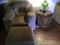 Photo showing brown upholstered armchair, matching foot stool, wooden carved hexagonal end table on hardwood floor