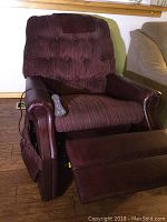 Burgundy recliner chair with plush tufted back and ribbed seating cushion. Armrests and outer sides covered in matching burgundy leather or faux leather showing wear. Massage control remote attached by cord is resting on the seat.