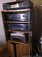 Photo of stereo components stacked on wooden audio stand with glass door, includes TDK CD player on top, Pioneer tape deck, receiver, and VCR below, with some vinyl or papers stored on one shelf.