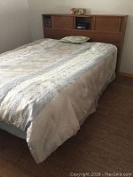 Double size bed with wooden headboard, mattress covered with light-colored bedding and pillow, hardwood floor visible