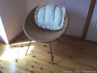 Photo showing a round wicker chair with thin black metal legs placed on wooden flooring. A cream and blue patterned cushion with ruffled edges sits on the chair.