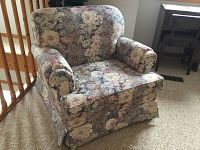 Floral patterned armchair with rolled armrests and skirted base on carpeted floor near wooden railing and dark wood side table.