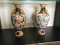 Pair of antique ceramic vases shown side by side on a black surface against a neutral background, showcasing their ornate handles and colorful floral and fruit pattern decoration.