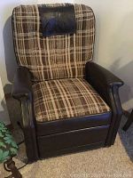 Front view of the reclining massage chair showing brown plaid upholstery and dark brown leather arms and base with attached black cushion headrest.