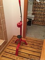 Red manual wine bottle corker with a long handle and tripod base placed on a wooden surface, against a wall.