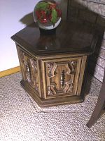 Side angle of a hexagonal wooden end table featuring carved wood panels and a small metal handle on the door.