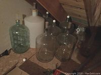 Image showing six clear glass carboys and one white plastic carboy arranged on a wooden floor under an attic roof. Some have stoppers or caps on top.